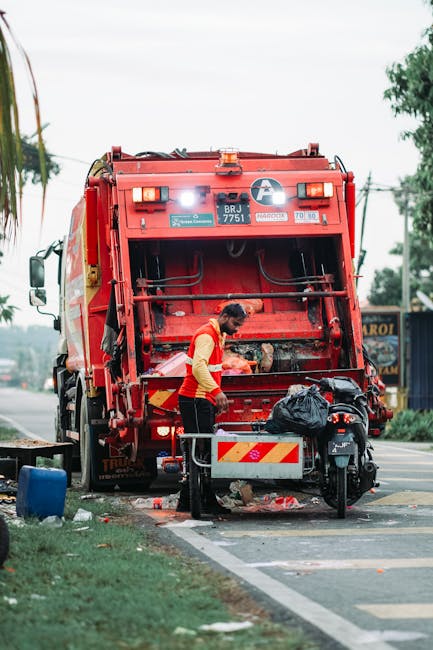 A waste collection truck, predominantly white with signs of rust and wear, is parked on a narrow urban street adjacent to aged residential buildings with weathered facades and multiple windows. The tr