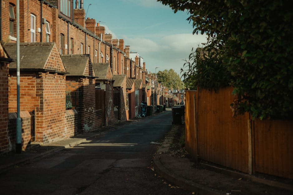 A narrow residential street lined with red brick terraced houses featuring small front gardens and peaked tiled roofs. The street pavement is paved with asphalt, with a small curb separating it from t