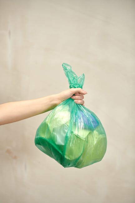 A person's hand is holding a green plastic bag filled with various types of waste materials, including what appears to be a blue plastic object, against a neutral beige background. The bag is tied at 